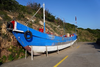 A large blue and white wooden boat is stationed on a paved pathway beside a rocky and grassy hillside. The boat is supported by wooden stands and has red accents, including a flag on top. There are black tires hanging on the side of the boat.