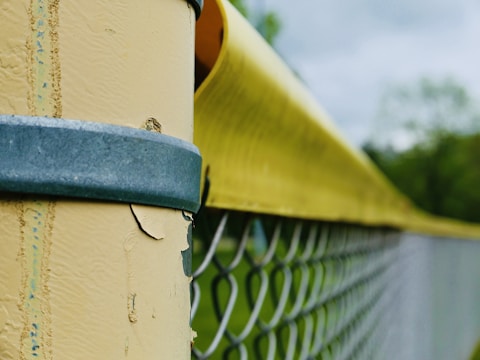 A close-up view of a fence post with peeling yellow paint, secured by a blue metal band. A chain-link fence extends into the background, with green foliage and a cloudy sky visible in the distance.