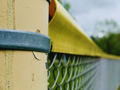 A close-up view of a fence post with peeling yellow paint, secured by a blue metal band. A chain-link fence extends into the background, with green foliage and a cloudy sky visible in the distance.