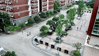 An aerial view of a residential area with multi-story red brick buildings and a paved plaza. The scene includes several trees, pedestrians walking along paths, and a few parked bicycles. The area is clean and well-maintained, with neatly arranged green spaces and street lamps.