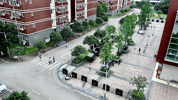 An aerial view of a residential area with multi-story red brick buildings and a paved plaza. The scene includes several trees, pedestrians walking along paths, and a few parked bicycles. The area is clean and well-maintained, with neatly arranged green spaces and street lamps.