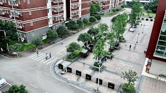An aerial view of a residential area with multi-story red brick buildings and a paved plaza. The scene includes several trees, pedestrians walking along paths, and a few parked bicycles. The area is clean and well-maintained, with neatly arranged green spaces and street lamps.