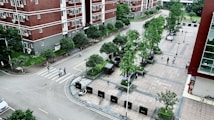 An aerial view of a residential area with multi-story red brick buildings and a paved plaza. The scene includes several trees, pedestrians walking along paths, and a few parked bicycles. The area is clean and well-maintained, with neatly arranged green spaces and street lamps.