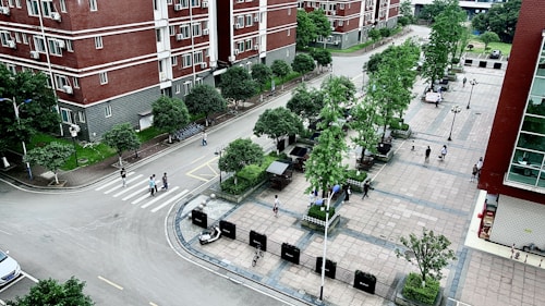 An aerial view of a residential area with multi-story red brick buildings and a paved plaza. The scene includes several trees, pedestrians walking along paths, and a few parked bicycles. The area is clean and well-maintained, with neatly arranged green spaces and street lamps.