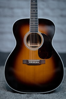 An acoustic guitar with a sunburst finish, prominently featuring the body and strings. The guitar has a glossy surface, showcasing a warm gradient from dark brown to light amber. It rests on a soft, textured surface against a blurred, neutral background.