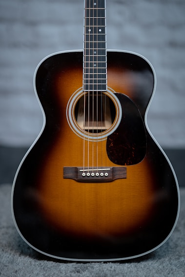 An acoustic guitar with a sunburst finish, prominently featuring the body and strings. The guitar has a glossy surface, showcasing a warm gradient from dark brown to light amber. It rests on a soft, textured surface against a blurred, neutral background.