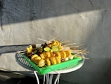 A green tray is placed on a round metal table, filled with skewers of corn, slices of cucumber, and other vegetables. Sunlight casts shadows across the gray textured wall in the background.