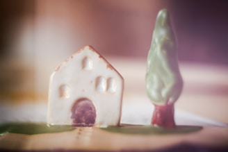 A small ceramic or clay model of a house with a slightly textured surface, featuring a rounded archway and several small windows. Next to the house is a model of a tree with a tall, tapered shape. The scene uses soft, warm lighting, creating a blurred background.