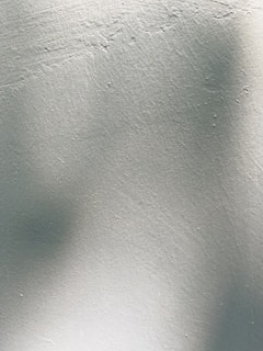 A textured surface of a light-colored wall with subtle shadows and unevenness. The texture suggests a rough, painted finish.