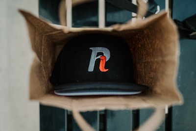 A brown paper bag holds a black baseball cap with a stylized letter logo featuring gray and red colors. The cap is positioned at the bottom of the bag, while the bag itself is situated on a striped surface.