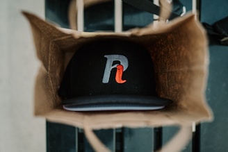 A brown paper bag holds a black baseball cap with a stylized letter logo featuring gray and red colors. The cap is positioned at the bottom of the bag, while the bag itself is situated on a striped surface.