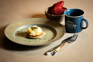 A plate featuring a well-prepared eggs Benedict topped with hollandaise sauce, garnished with herbs, accompanied by a set of silver fork and knife. Next to the plate is a blue mug with 'Adventure Awaits' written on it, filled with a dark beverage. A wooden bowl containing fresh strawberries is placed beside the mug.
