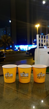 Three yellow cups with white rims and 'Chai Street' written on them are placed on a glass table. In the background, there is a night street scene with blurred lights from buildings and vehicles. A white railing and some parked cars are also visible.