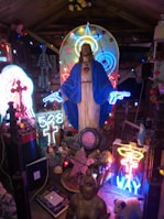 A vibrant religious-themed display featuring a statue of Jesus surrounded by colorful neon lights and eclectic items. The statue is adorned with a blue robe and Sacred Heart imagery, accented by neon signs shaped like guns, stars, and crosses. Various other objects, including a buddha sculpture, notebooks, and a small dog plush, are placed around the statue. The setting is a wooden room adorned with additional lights and flower decorations.