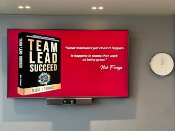 A large screen displays a book titled 'Team Lead Succeed' by Nick Fewings next to a motivational quote about teamwork attributed to the same author. The background is predominantly red. To the right of the screen, there is a white analog clock. Below the screen, a video conferencing camera is mounted.