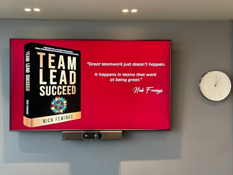 A large screen displays a book titled 'Team Lead Succeed' by Nick Fewings next to a motivational quote about teamwork attributed to the same author. The background is predominantly red. To the right of the screen, there is a white analog clock. Below the screen, a video conferencing camera is mounted.
