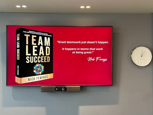 A large screen displays a book titled 'Team Lead Succeed' by Nick Fewings next to a motivational quote about teamwork attributed to the same author. The background is predominantly red. To the right of the screen, there is a white analog clock. Below the screen, a video conferencing camera is mounted.