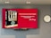 A large screen displays a book titled 'Team Lead Succeed' by Nick Fewings next to a motivational quote about teamwork attributed to the same author. The background is predominantly red. To the right of the screen, there is a white analog clock. Below the screen, a video conferencing camera is mounted.