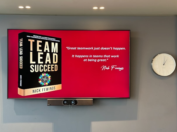 A large screen displays a book titled 'Team Lead Succeed' by Nick Fewings next to a motivational quote about teamwork attributed to the same author. The background is predominantly red. To the right of the screen, there is a white analog clock. Below the screen, a video conferencing camera is mounted.