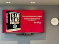 A large screen displays a book titled 'Team Lead Succeed' by Nick Fewings next to a motivational quote about teamwork attributed to the same author. The background is predominantly red. To the right of the screen, there is a white analog clock. Below the screen, a video conferencing camera is mounted.