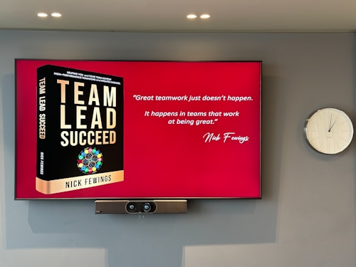 A large screen displays a book titled 'Team Lead Succeed' by Nick Fewings next to a motivational quote about teamwork attributed to the same author. The background is predominantly red. To the right of the screen, there is a white analog clock. Below the screen, a video conferencing camera is mounted.