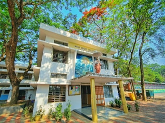 A two-story building surrounded by lush green trees and a clear blue sky. The front features a statue of a religious figure holding an umbrella, with signage reading 'Holy Cross College'. Several potted plants line the entrance, and a bulletin board is visible.