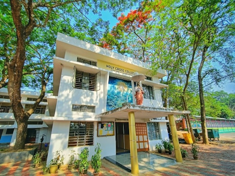 A two-story building surrounded by lush green trees and a clear blue sky. The front features a statue of a religious figure holding an umbrella, with signage reading 'Holy Cross College'. Several potted plants line the entrance, and a bulletin board is visible.