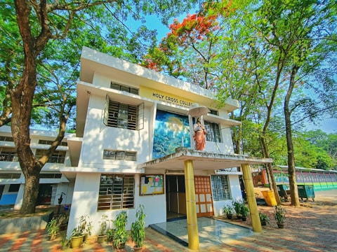 A two-story building surrounded by lush green trees and a clear blue sky. The front features a statue of a religious figure holding an umbrella, with signage reading 'Holy Cross College'. Several potted plants line the entrance, and a bulletin board is visible.