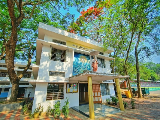 A two-story building surrounded by lush green trees and a clear blue sky. The front features a statue of a religious figure holding an umbrella, with signage reading 'Holy Cross College'. Several potted plants line the entrance, and a bulletin board is visible.