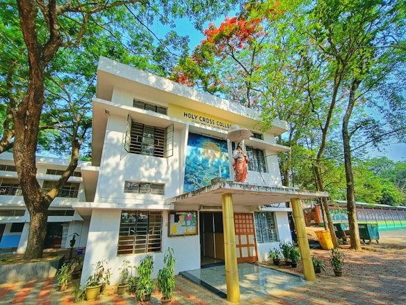 A two-story building surrounded by lush green trees and a clear blue sky. The front features a statue of a religious figure holding an umbrella, with signage reading 'Holy Cross College'. Several potted plants line the entrance, and a bulletin board is visible.