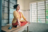 A person is meditating in a seated cross-legged position on a wooden table in a room with barred windows, wearing a yellow tunic and red pants. A smartphone is placed beside them on the table.