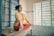 A person is meditating in a seated cross-legged position on a wooden table in a room with barred windows, wearing a yellow tunic and red pants. A smartphone is placed beside them on the table.