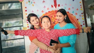 Three women are standing close together, smiling joyfully. The woman in the center is holding her arms wide open and wearing glasses and a red patterned shirt. Behind them is a colorful wall decorated with various cut-out shapes and a large orange semi-circle design with a red arrow pointing upwards.