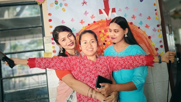 Three women are standing close together, smiling joyfully. The woman in the center is holding her arms wide open and wearing glasses and a red patterned shirt. Behind them is a colorful wall decorated with various cut-out shapes and a large orange semi-circle design with a red arrow pointing upwards.