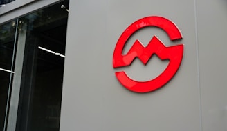 A red abstract logo is mounted on a large, smooth gray surface next to a glass wall, which appears to be part of a modern building facade.