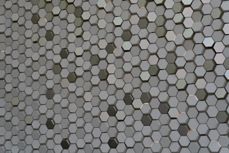 A pattern of countless hexagonal tiles creating a geometric texture. These tiles have a three-dimensional appearance with varying shades of grey and hints of subtle reflections, giving them a metallic or ceramic look.