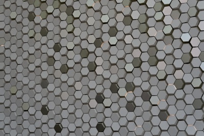 A pattern of countless hexagonal tiles creating a geometric texture. These tiles have a three-dimensional appearance with varying shades of grey and hints of subtle reflections, giving them a metallic or ceramic look.