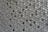 A pattern of countless hexagonal tiles creating a geometric texture. These tiles have a three-dimensional appearance with varying shades of grey and hints of subtle reflections, giving them a metallic or ceramic look.