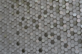 A pattern of countless hexagonal tiles creating a geometric texture. These tiles have a three-dimensional appearance with varying shades of grey and hints of subtle reflections, giving them a metallic or ceramic look.