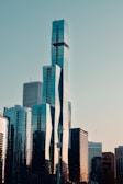 A skyline featuring a cluster of modern skyscrapers with unique architectural designs, reflecting the sunset light. The buildings are composed of glass and steel, and a clear sky serves as the backdrop, with a single bird in flight.