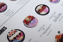 A close-up of a menu displaying various smoothie bowls. The bowls are placed in rows with descriptions and calorie counts next to each. The bowls are decorated with fresh fruits like strawberries, blueberries, and exotic fruits. The design and layout are clean with a white background and aesthetically pleasing presentation.