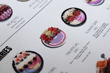 A close-up of a menu displaying various smoothie bowls. The bowls are placed in rows with descriptions and calorie counts next to each. The bowls are decorated with fresh fruits like strawberries, blueberries, and exotic fruits. The design and layout are clean with a white background and aesthetically pleasing presentation.