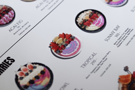 A close-up of a menu displaying various smoothie bowls. The bowls are placed in rows with descriptions and calorie counts next to each. The bowls are decorated with fresh fruits like strawberries, blueberries, and exotic fruits. The design and layout are clean with a white background and aesthetically pleasing presentation.