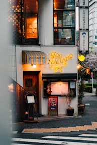 A cozy Italian restaurant located on a street corner. The entrance features a wooden door next to a yellow neon sign displaying 'Goo Italiano'. Menu boards and small plants are positioned outside, with warm light emanating from windows above showing decorative elements inside. There's a striped awning above one of the windows.