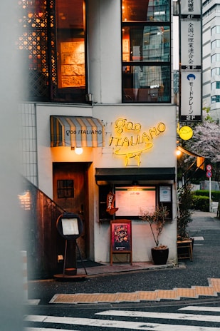 A cozy Italian restaurant located on a street corner. The entrance features a wooden door next to a yellow neon sign displaying 'Goo Italiano'. Menu boards and small plants are positioned outside, with warm light emanating from windows above showing decorative elements inside. There's a striped awning above one of the windows.