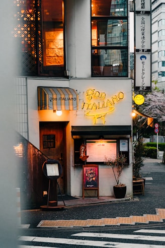 A cozy Italian restaurant located on a street corner. The entrance features a wooden door next to a yellow neon sign displaying 'Goo Italiano'. Menu boards and small plants are positioned outside, with warm light emanating from windows above showing decorative elements inside. There's a striped awning above one of the windows.