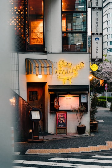 A cozy Italian restaurant located on a street corner. The entrance features a wooden door next to a yellow neon sign displaying 'Goo Italiano'. Menu boards and small plants are positioned outside, with warm light emanating from windows above showing decorative elements inside. There's a striped awning above one of the windows.