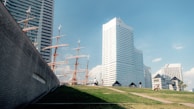 A landscape featuring modern high-rise buildings with unique designs adjacent to a sailing ship with multiple masts. The foreground includes a grassy area with pathways and a few people walking or sitting, contributing to a serene urban setting under a clear blue sky.