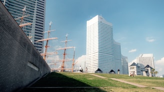 A landscape featuring modern high-rise buildings with unique designs adjacent to a sailing ship with multiple masts. The foreground includes a grassy area with pathways and a few people walking or sitting, contributing to a serene urban setting under a clear blue sky.