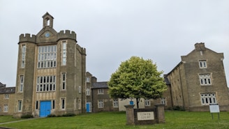 An old, castle-like building with a prominent central tower featuring a round clock near the top. The structure is constructed from grayish stone and has several windows with white frames. The central section has a blue door at the bottom. A lush green tree stands in front of the building, and a sign reading 'Tower Court' is positioned near the tree.